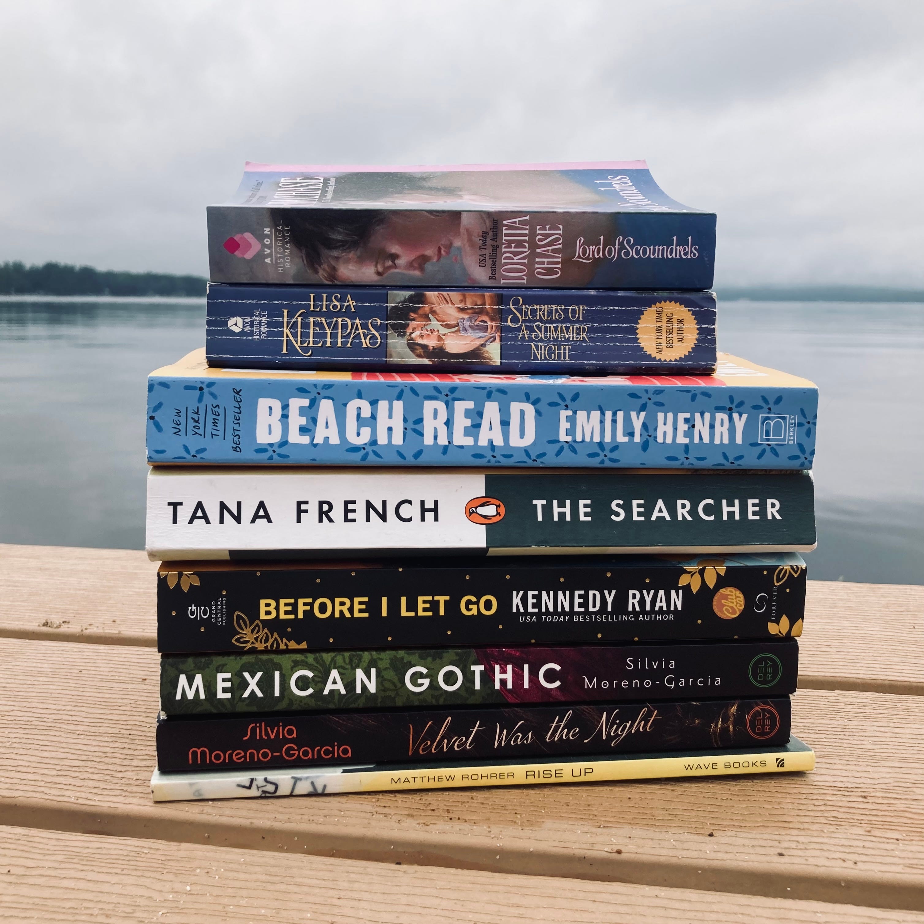 eight paperback books (named and described in the text below) stacked on the end of a dock with the view of a lake in the background