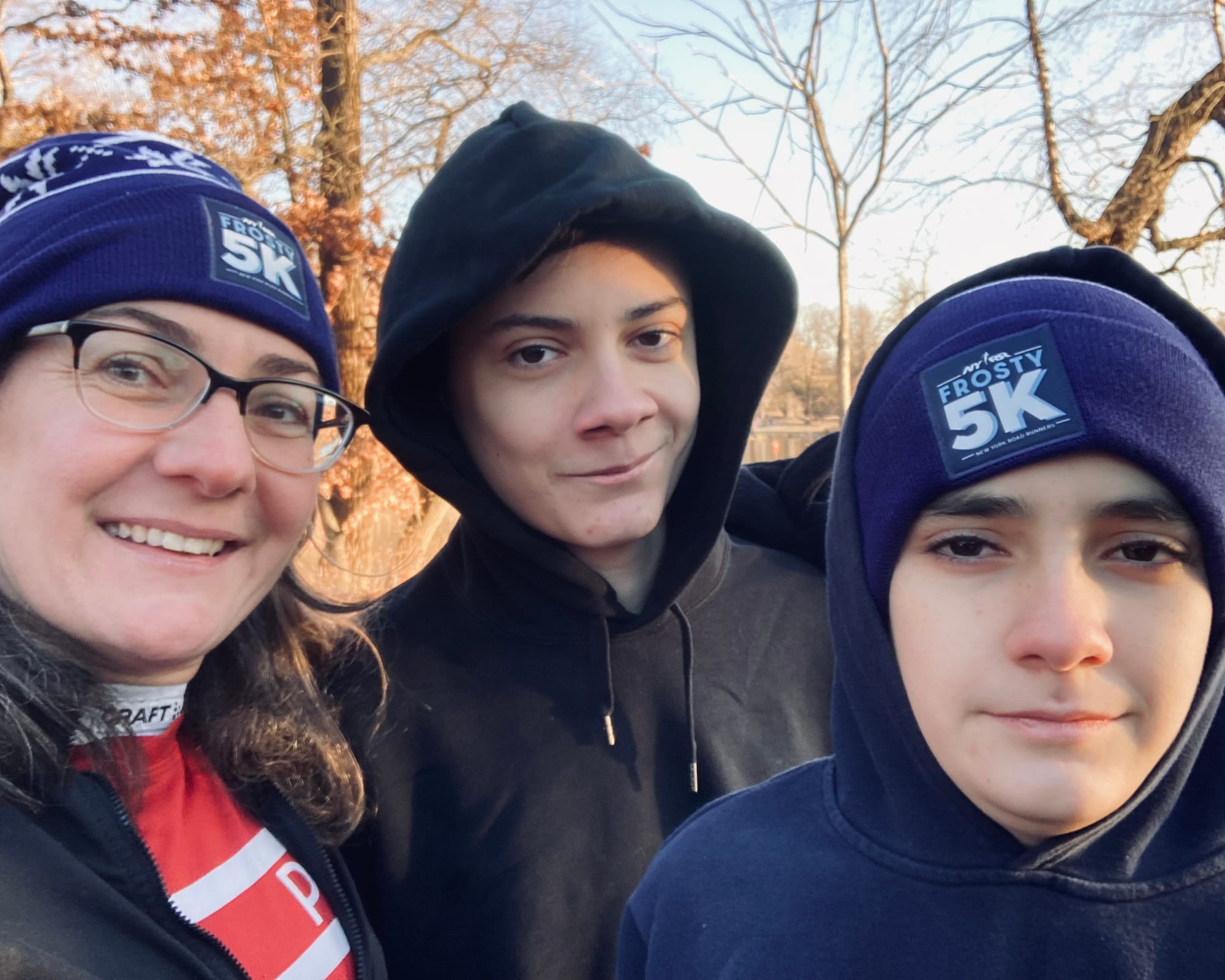 a selfie with my two children, the older one smiling and the younger one not, with bare trees and the Prospect Park Lake behind us