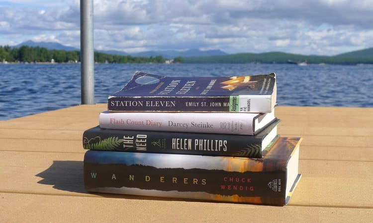 books (Station 11, Flash Count Diary, The Need, Wanderers) stacked on the edge of a dock with the view of a lake & mountains