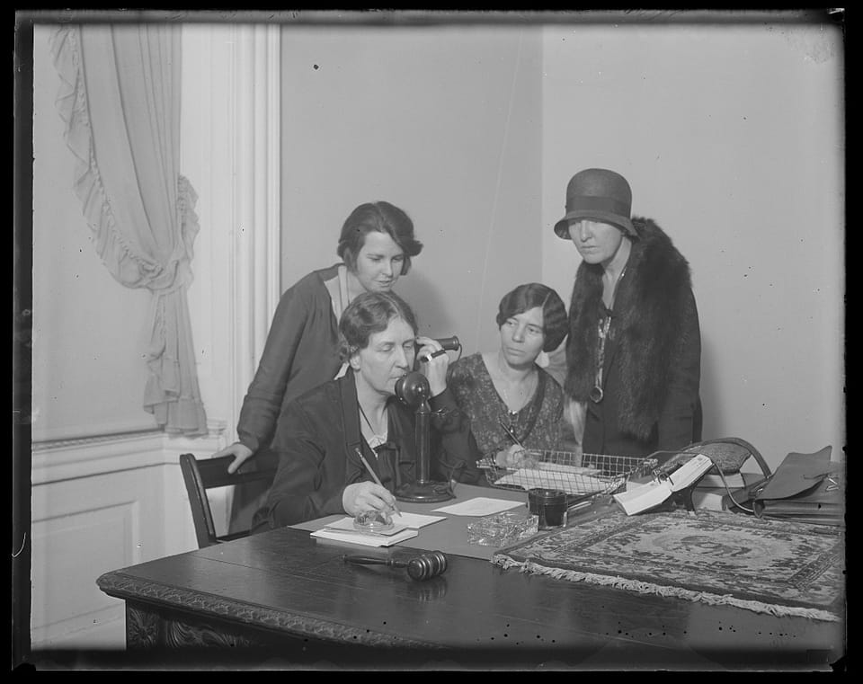 black-and-white photo showing 3 women in 1930s dress sitting or standing around another woman on the telephone