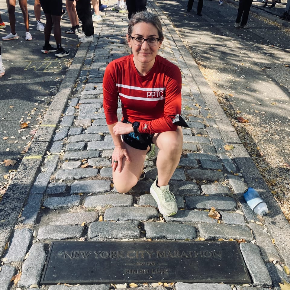 A runner smiling and half squatting, half kneeling beside a plaque that says New York City Marathon Est. 1970 Finish Line